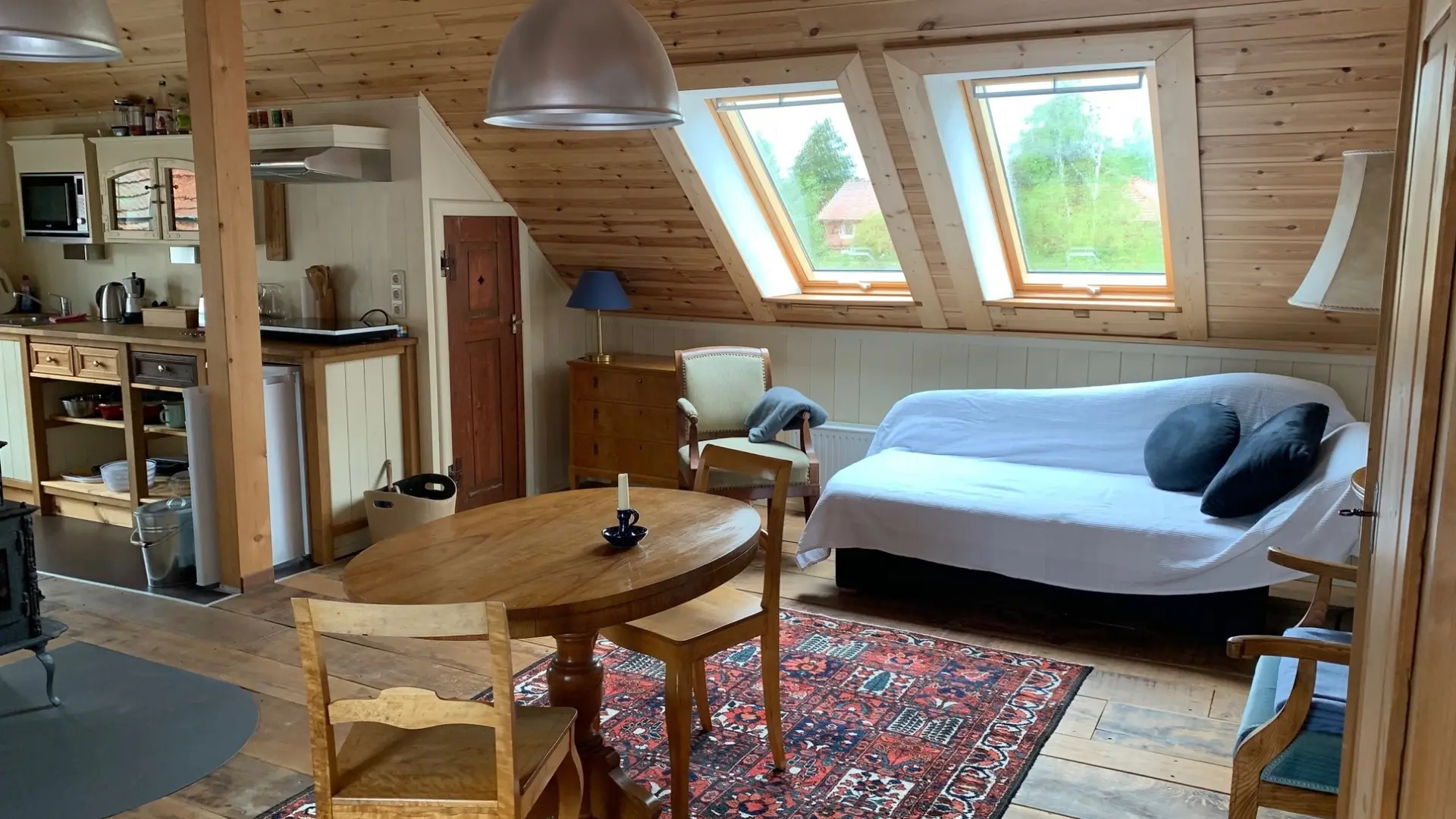 Sicht in den Wohnbereich. Zu sehen ist das Sofa, welches als Einzelbett genutzt werden kann, ein ovaler Tisch vor dem Ofen, eine Kommode und Einblick in die Küchenzeile 